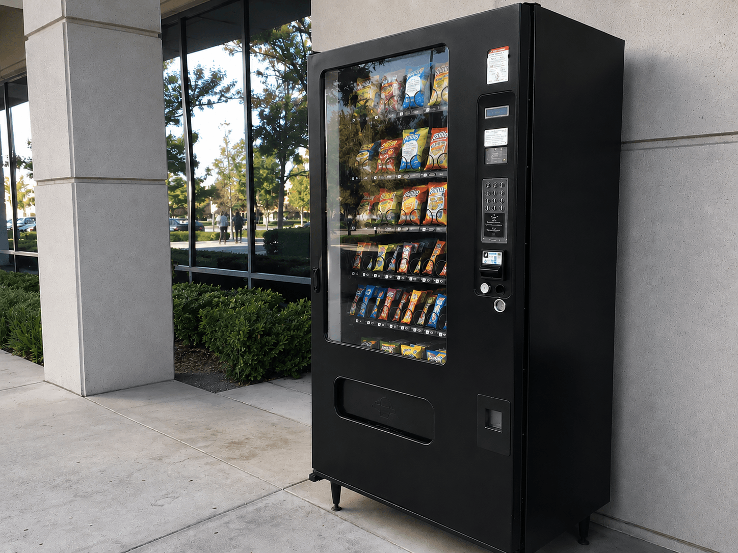 A black snack vending machine outside a modern office building, with landscaped greenery, reflective windows, and a couple of people walking in the background.